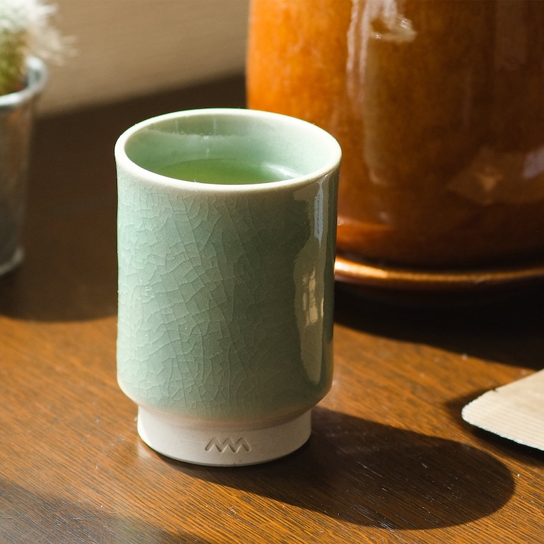 Op de afbeelding: Een lichtgroene keramische theekop met een craquel&eacute; glazuur, gevuld met groene thee. De beker heeft een witte basis met een gestileerd 'M'-ontwerp. De beker staat op een houten oppervlak, met een vage achtergrond.