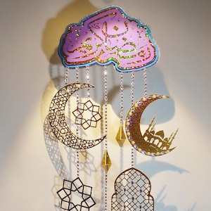 May include: A gold and purple Ramadan decoration with a cloud-shaped top featuring Arabic calligraphy. The decoration hangs from a string with several gold and silver ornaments, including crescent moons, stars, and a lantern.