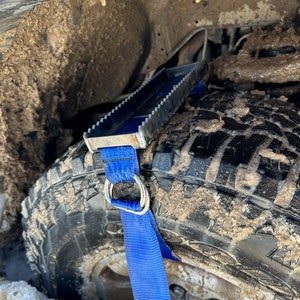May include: A blue strap with a metal loop is attached to a metal traction device. The device is placed under a muddy tire.