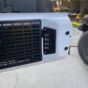 May include: A white vehicle with a black metal grate and a black plastic panel with multiple slots. The vehicle is parked on a gray concrete surface.
