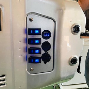May include: A silver panel featuring four blue-lit switches, a digital voltage display showing 12.3 volts, and two DC power outlets. The panel is mounted on a larger silver surface.