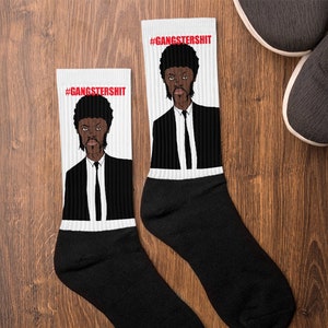 May include: A pair of black socks with a white background featuring a cartoon image of a man with a black afro wearing a black suit and a white tie. The text '#GANGSTERSHIT' is printed in red at the top of each sock.