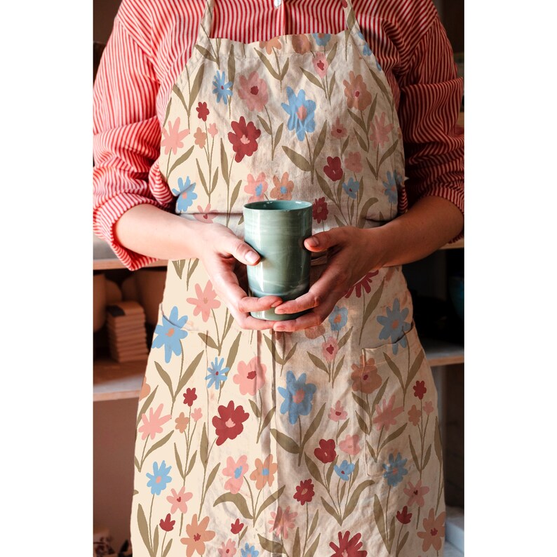 Puede incluir: Un delantal beige con estampado floral con flores rosas, azules y rojas. El delantal tiene un bolsillo grande en la parte delantera.