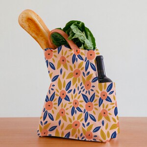 Pode incluir: Uma bolsa de tecido reutiliz&aacute;vel com um padr&atilde;o floral em tons de rosa, azul e amarelo. A bolsa tem uma al&ccedil;a de couro marrom e est&aacute; cheia de um p&atilde;o, um molho de vegetais de folhas verdes e uma garrafa de vinho.