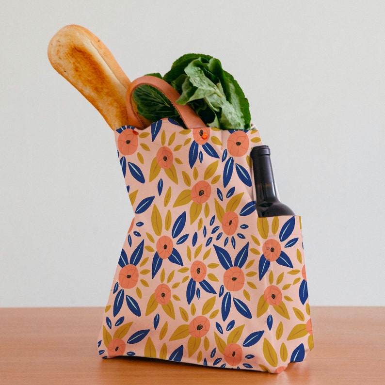 Pode incluir: Uma bolsa de tecido reutiliz&aacute;vel com um padr&atilde;o floral em tons de rosa, azul e amarelo. A bolsa tem uma al&ccedil;a de couro marrom e est&aacute; cheia de um p&atilde;o, um molho de vegetais de folhas verdes e uma garrafa de vinho.