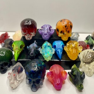 May include: A collection of colorful resin skull sculptures in various sizes and styles. The skulls are in shades of red, purple, blue, green, and yellow. Some have a glossy finish, while others are translucent. The skulls are displayed on a white surface.
