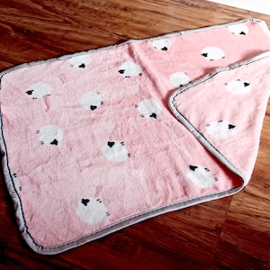 Könnte beinhalten: Eine rosa Fleece-Decke mit einem weißen Schaf-Design und einem grauen Saum.