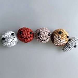 Puede incluir: Seis caras sonrientes de ganchillo en varios colores, incluyendo gris, rojo y beige. Cada cara tiene ojos de botón negros y una sonrisa cosida. Las caras están dispuestas sobre un fondo gris claro.