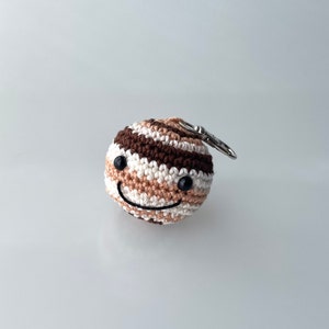 Peut inclure: Une boule en crochet de couleur marron, blanche et beige avec un visage souriant et des yeux noirs. La boule est attachée à un porte-clés argenté.