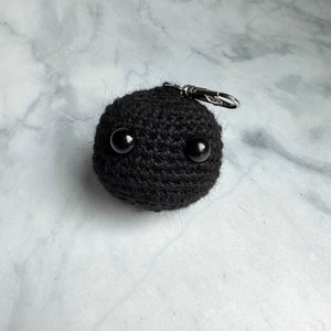 Puede incluir: Llavero de crochet negro con dos ojos de botón negros y un cierre de metal plateado.