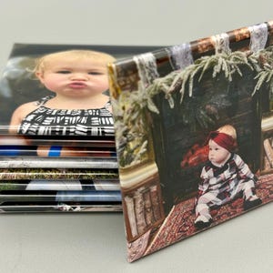 May include: A stack of square photo magnets with images of children. The top magnet features a young girl with a black and white patterned shirt. The bottom magnet features a young girl in a plaid dress sitting in front of a fireplace.