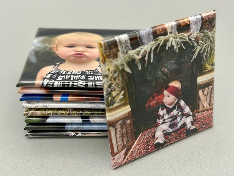 May include: A stack of square photo magnets with images of children. The top magnet features a young girl with a black and white patterned shirt. The bottom magnet features a young girl in a plaid dress sitting in front of a fireplace.