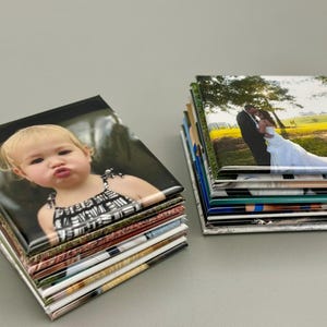 May include: A stack of square photo magnets with a variety of images, including a baby girl and a wedding couple. The magnets are arranged in a neat pile on a gray surface.