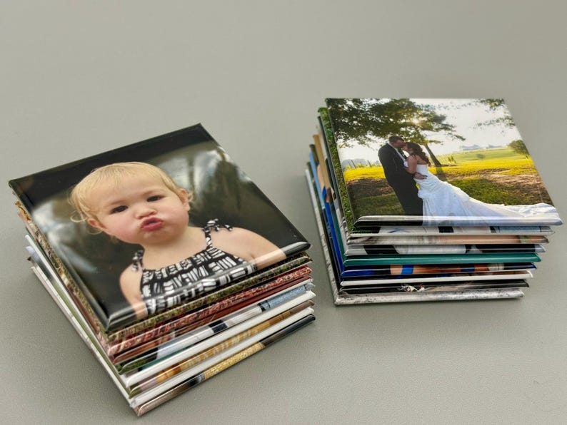 May include: A stack of square photo magnets with a variety of images, including a baby girl and a wedding couple. The magnets are arranged in a neat pile on a gray surface.