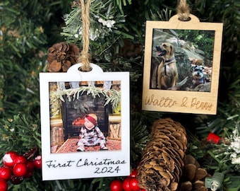Puede incluir: Dos adornos de fotos de madera y blanco con una cuerda para colgar. El primer adorno tiene una foto de un bebé con un atuendo a cuadros sentado frente a una chimenea. El texto en el adorno dice "First Christmas 2024". El segundo adorno tiene una foto de dos perros, uno marrón y uno gris, de pie en un área boscosa. El texto en el adorno dice "Walter & Penny".