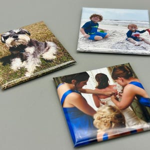 May include: Three square photo magnets with different images. The first magnet features a small black and gray dog sitting on green grass. The second magnet shows two children on a beach. The third magnet shows three people in blue dresses getting ready for a wedding.
