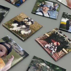 May include: A collection of square photo magnets with various images, including a family portrait, a dog, a baby, and a couple. The magnets are arranged on a gray surface.