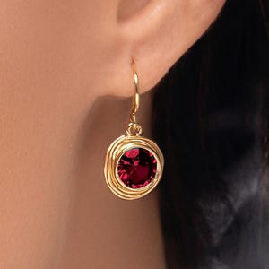 May include: Gold-tone drop earring featuring a round, deep red gemstone. The gemstone is set within a spiral design. The earring is shown worn on an ear.