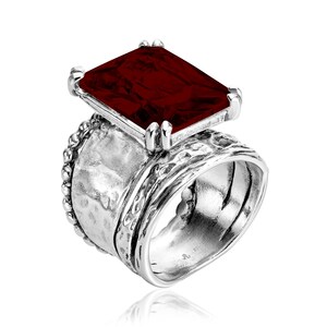 May include: A silver ring with a large, dark red, faceted gemstone. The ring band is textured and has a beaded edge.