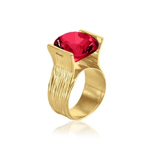 May include: A gold-colored ring featuring a large, faceted, red gemstone. The ring has a textured, organic design with a wide band and decorative elements that cradle the gemstone. The ring is set against a white background.