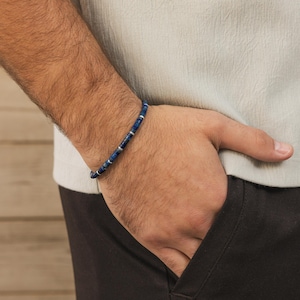 May include: A blue beaded bracelet with silver-colored spacers. The bracelet is worn on a wrist, with the hand resting in a pocket. The beads appear to be square or rectangular in shape, and the bracelet is a simple, elegant design.