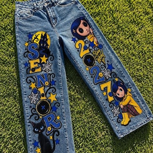 May include: Blue denim jeans featuring custom painted graphics. The left leg displays the word SENIOR in blue and yellow with a black cat and a haunted house. The right leg features the text 2027, Stay Weird, and two cartoon characters in yellow raincoats.