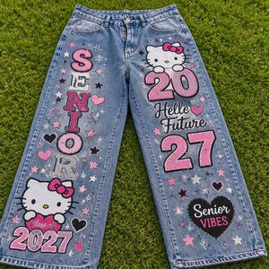 Puede incluir: Vaqueros de mezclilla azul claro con gráficos personalizados que incluyen a Hello Kitty, corazones rosas y negros, y estrellas. El texto dice SENIOR, 20, Hello Future, 27, Class of 2027 y Senior Vibes. El diseño incorpora detalles en rosa y plata.