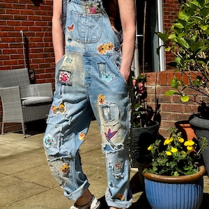 Può includere: Salopette in denim azzurro con finitura consumata, toppe sfilacciate e varie applicazioni ricamate colorate tra cui farfalle, fiori e un colibrì. Il capo presenta una tasca sul pettorale e i bordi dei pantaloni risvoltati.