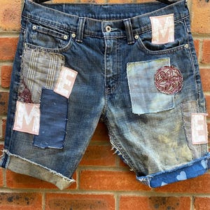May include: Denim shorts in blue with patchwork detailing. Various fabric patches are sewn onto the shorts, including an embroidered circular patch. The shorts have rolled cuffs, and the letters "M" and "E" are visible on some patches.