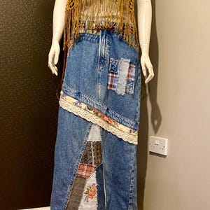 May include: A mannequin showcases a bohemian outfit. It features a mustard crochet top with fringe, a denim skirt with patchwork, and a floral ruffle hem. A layered necklace completes the look. The skirt is approximately 100 cm in length.