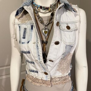 May include: A light-wash denim vest with patches and lace trim. The vest features brown buttons and is layered with multiple necklaces. The mannequin is also wearing a brown belt and blue jeans.
