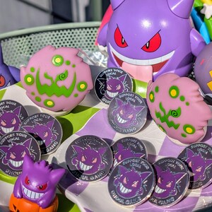 May include: A collection of 10 silver pins featuring a purple cartoon ghost with the text "Support Your Local Ghoul Gang". The pins are arranged on a purple and white checkered surface with a pink and green ghost figurine in the background.