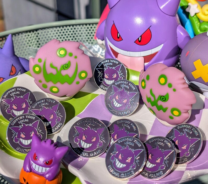 May include: A collection of 10 silver pins featuring a purple cartoon ghost with the text "Support Your Local Ghoul Gang". The pins are arranged on a purple and white checkered surface with a pink and green ghost figurine in the background.