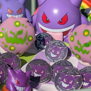 May include: A collection of enamel pins featuring a purple ghost character with a toothy grin. The pins are circular and have a white background with the text "Support Your Local Ghoul Gang".