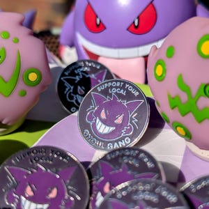May include: A close-up of a set of enamel pins featuring a purple cartoon ghost with a toothy grin. The pins are silver with black outlines and the text "Support Your Local Ghoul Gang".