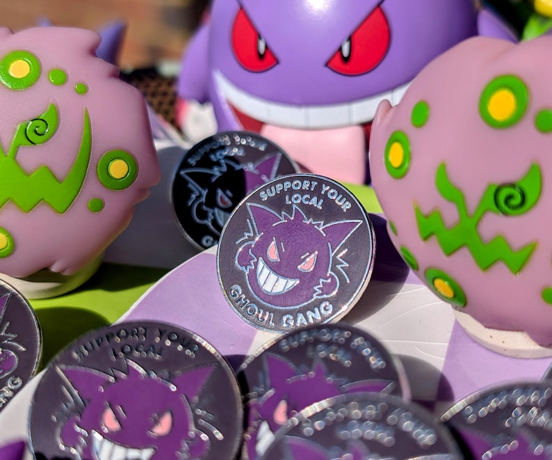 May include: A close-up of a set of enamel pins featuring a purple cartoon ghost with a toothy grin. The pins are silver with black outlines and the text "Support Your Local Ghoul Gang".