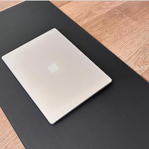 Pode incluir: Um tapete de mesa de couro preto com um computador portátil prateado em cima. O tapete está sobre uma superfície de madeira.