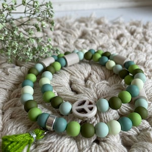 May include: Two layered bracelets with green, blue and white beads. The top layer has a white peace sign charm. The bracelets have a green tassel.