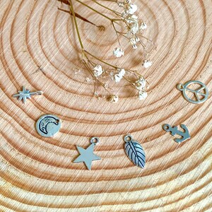 May include: A set of seven silver charms with various designs, including a star, crescent moon, peace sign, anchor, leaf, and two stars. The charms are displayed on a wooden surface.