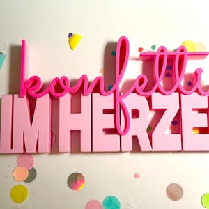Könnte beinhalten: Ein rosa Schild mit den Worten "Konfetti im Herzen". Das Wort "Konfetti" ist in einem leuchtenden Pink geschrieben, während "IM HERZEN" in einem blockartigen, hellrosa Schriftzug gehalten ist. Bunter Konfetti bedeckt den Hintergrund.