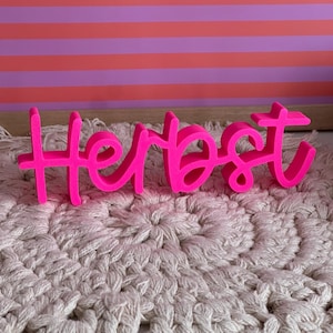Könnte beinhalten: Ein leuchtend neonpinkfarbenes 3D-Schild mit dem Wort Herbst in einer Schreibschrift. Die Buchstaben sind miteinander verbunden und stehen aufrecht, wobei sie eine kräftige, einfarbige Oberfläche aufweisen, die sich von der beigen Häkeloberfläche abhebt.