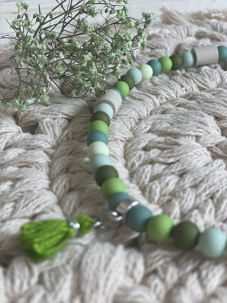 May include: A green and white beaded necklace with a green tassel charm. The necklace is made of silicone beads and is strung on a silver chain.