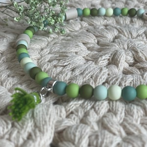 May include: A green and white beaded necklace with a green tassel charm. The necklace is on a white crocheted surface.
