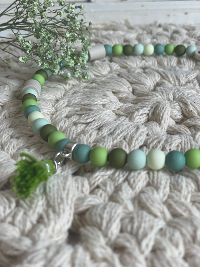 May include: A green and white beaded necklace with a green tassel charm. The necklace is on a white crocheted surface.