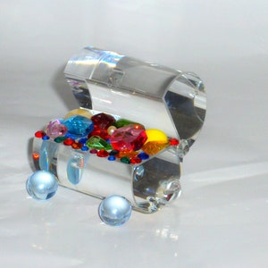 May include: A clear glass treasure chest filled with colorful gems and beads. The chest is open and the gems are visible. The chest is sitting on a white surface.
