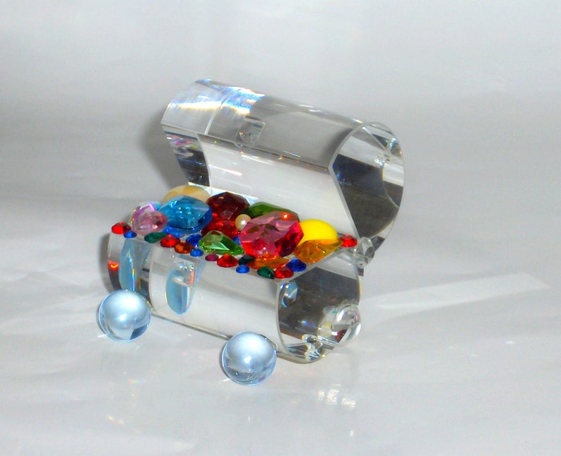 May include: A clear glass treasure chest filled with colorful gems and beads. The chest is open and the gems are visible. The chest is sitting on a white surface.