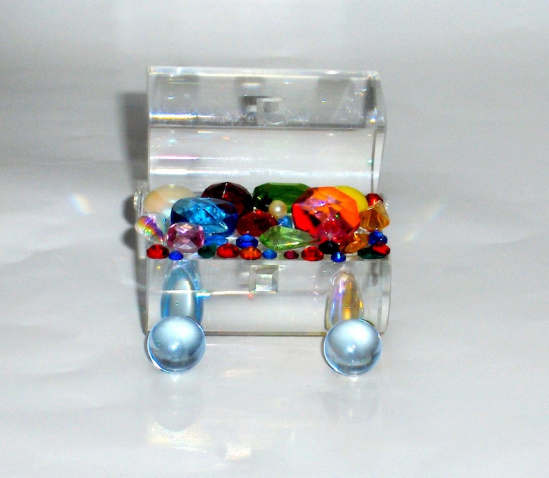 May include: A clear acrylic treasure chest filled with colorful jewels and beads. The chest has two round clear acrylic knobs on the front.