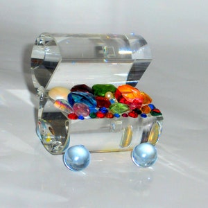 May include: A clear glass treasure chest filled with colorful gems and beads. The chest is on a white surface and has two clear glass wheels.