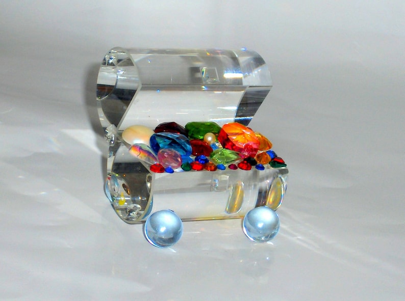 May include: A clear glass treasure chest filled with colorful gems and beads. The chest is on a white surface and has two clear glass wheels.