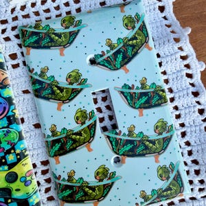 May include: Light blue light switch cover featuring a repeating pattern of a green creature relaxing in a bathtub with a rubber duck. The creature has a fish-like head and is surrounded by green plants. The cover has two screw holes and a switch opening.